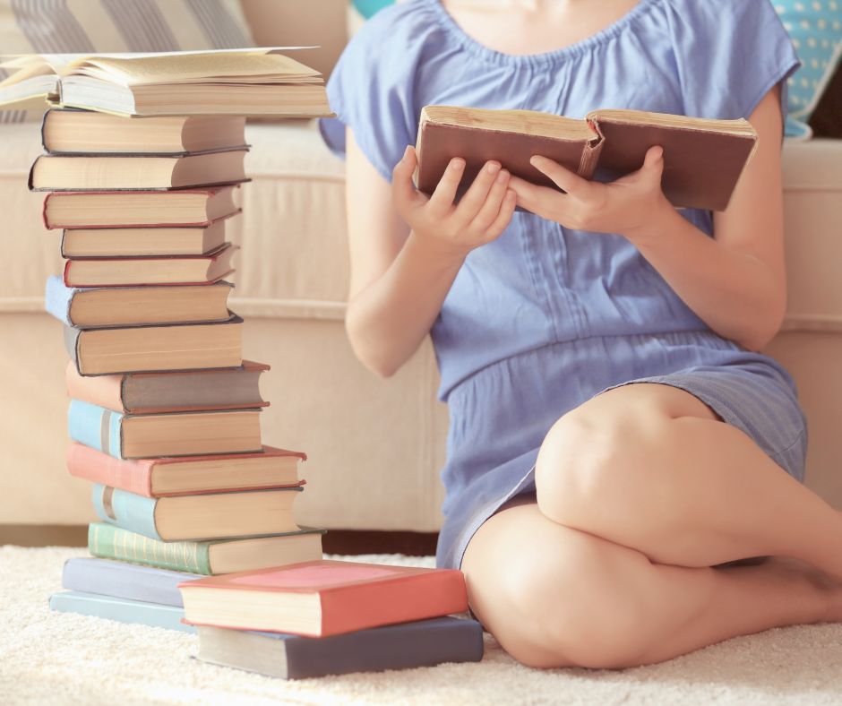 Image of a young woman in a blue dress sitting on the floor reading an old book with a large stack of books next to her.