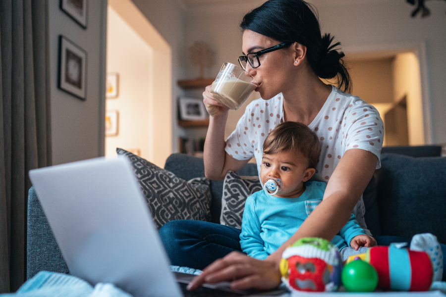 Woman sitting on a couch, using a laptop, while drinking coffee and holding a baby. If this is you, then join our Facebook Community for female sole providers, The Bright Side!